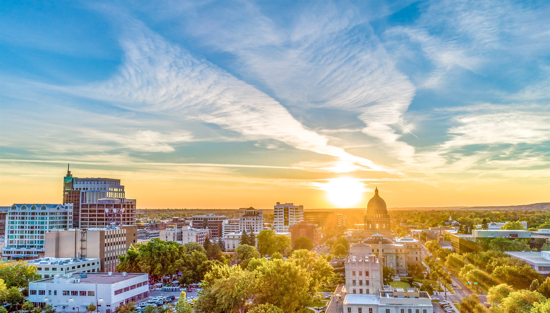 Downtown Boise at sunset under a wide cirrus sky, the sun dropping behind the Idaho State Capitol dome.
